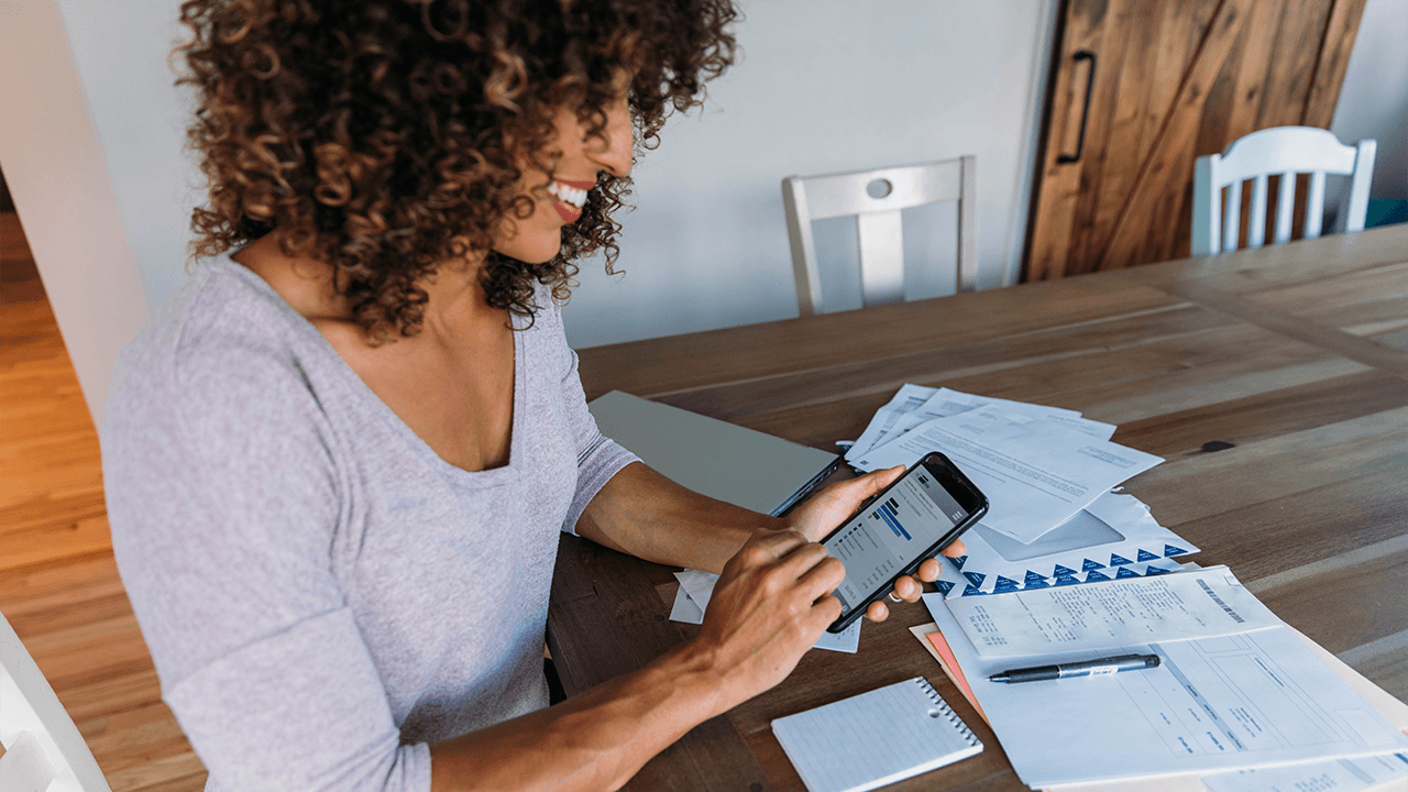 Mulher sorri enquanto checa a rentabilidade de seus investimentos na tela do celular